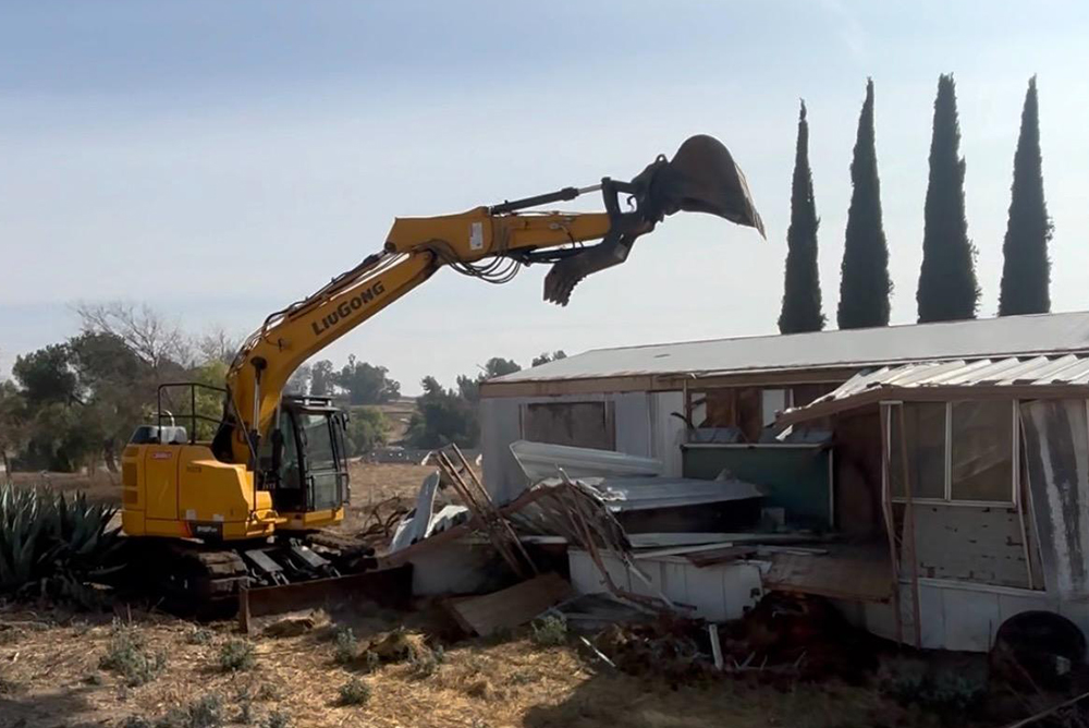 excavator performing total demolition of mobile home with cypress trees in background